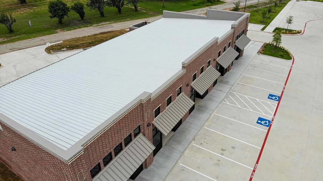 Aerial view of a brick retail building with a metal roof and awnings beside a striped concrete parking lot.