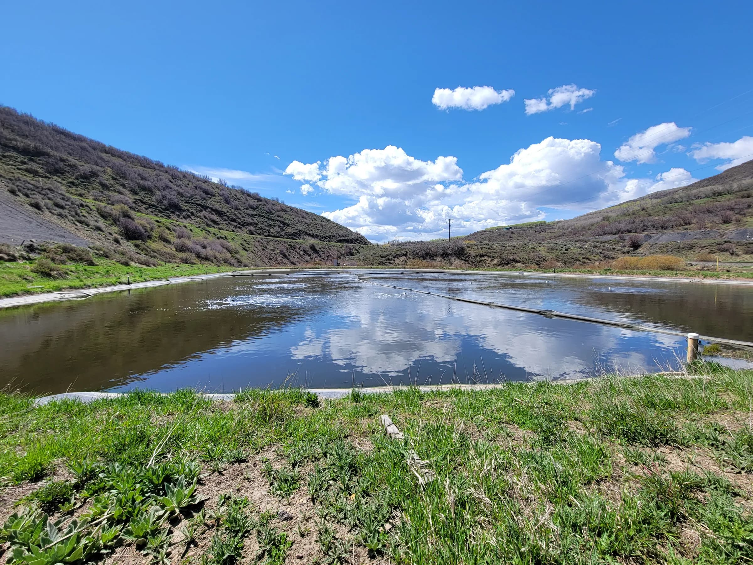 A lined, aerated wastewater lagoon in a green hillside valley under a blue sky with clouds.