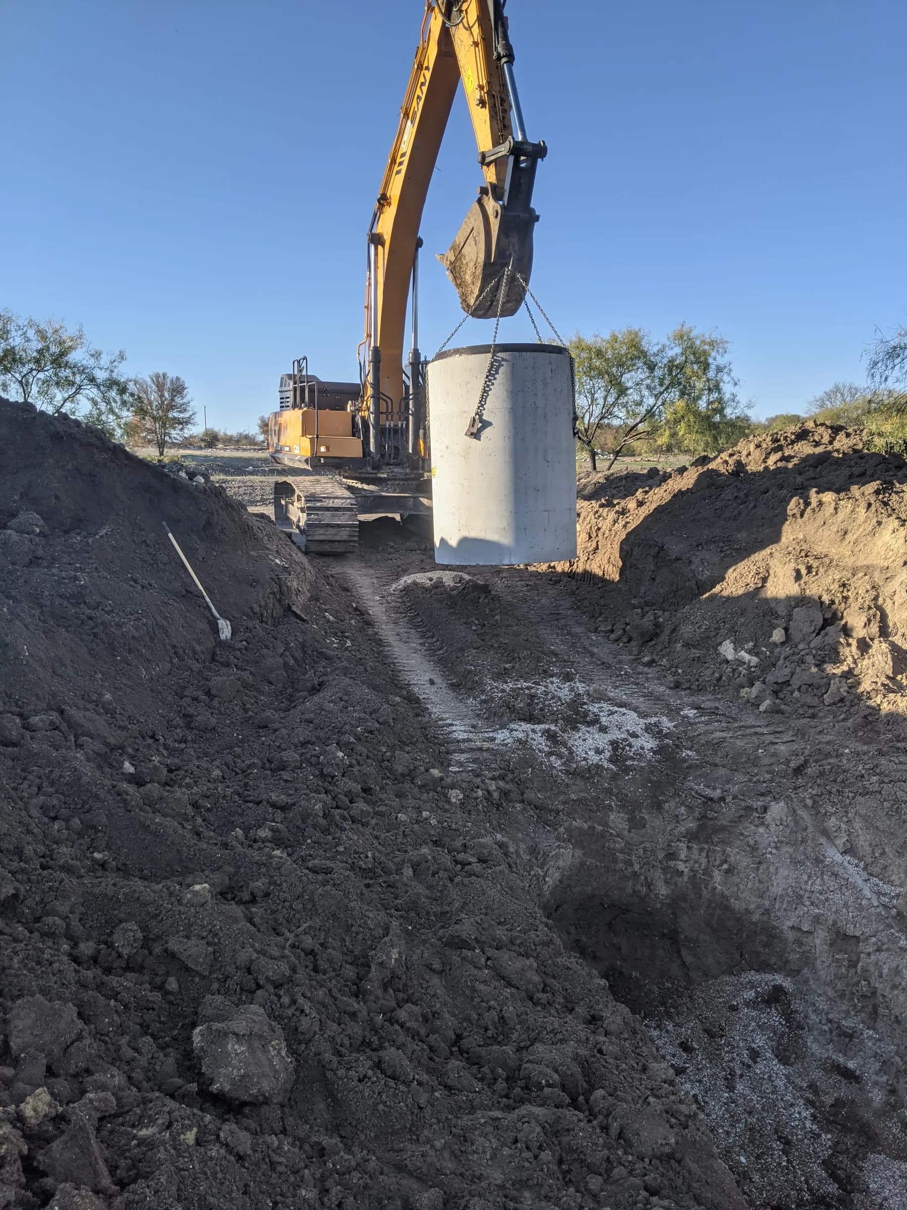 Tracked excavator lifting a large precast concrete lift station section on chains over an open excavation