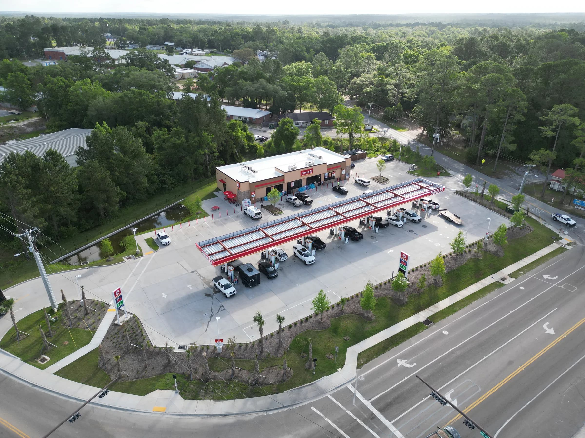 Aerial view of a convenience store and fuel station with a long canopy, a paved lot, a landscaped frontage and a drainage swale beside a divided highway, with pine woods behind.
