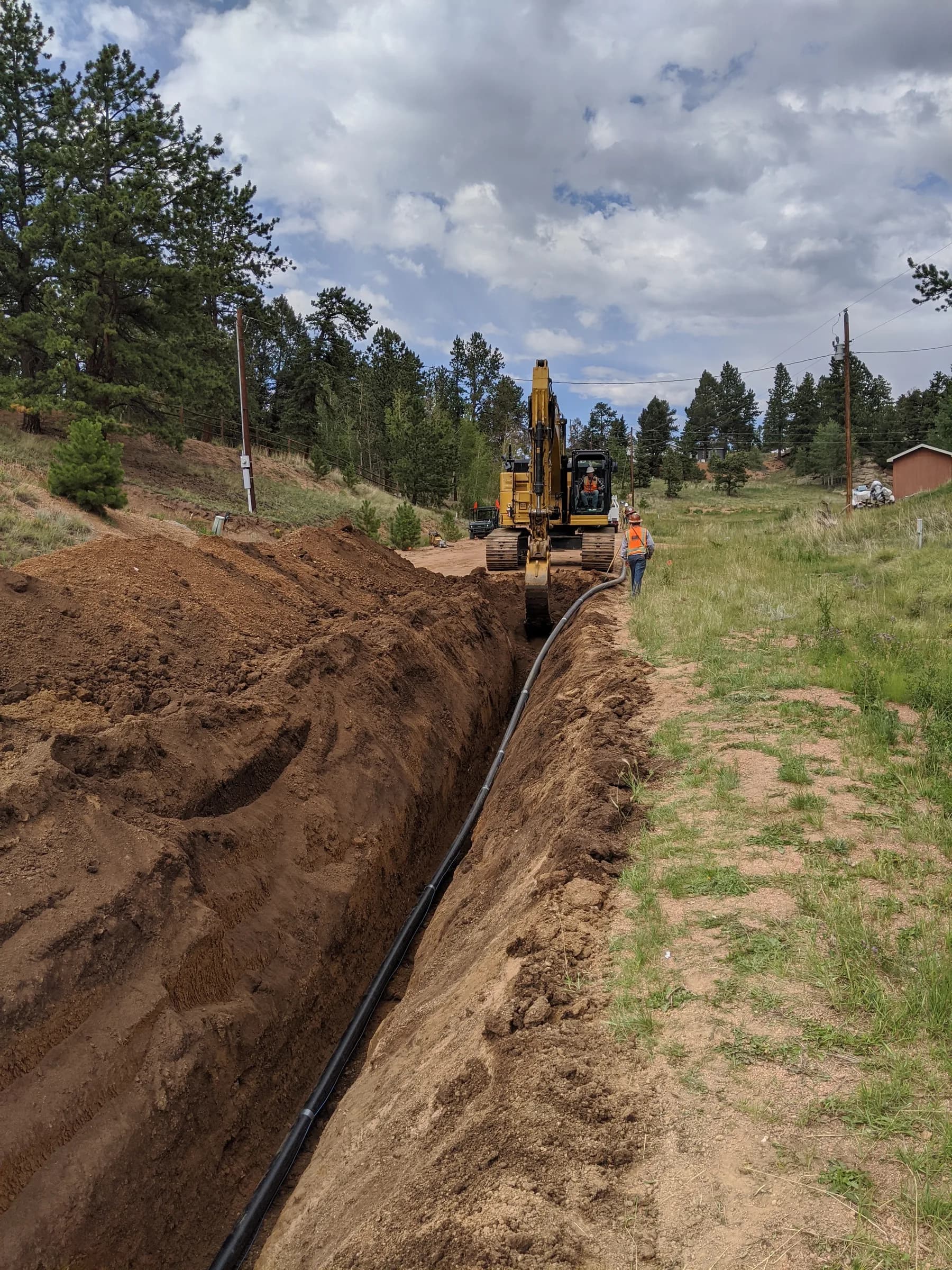 An excavator lays black HDPE pipe into a long open trench along a grassy right-of-way, with pines behind and a crew member watching.