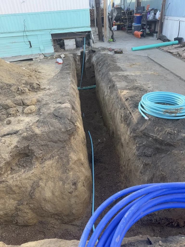 Open trench with new blue water pipe beside a mobile home, with coils of pipe on the ground