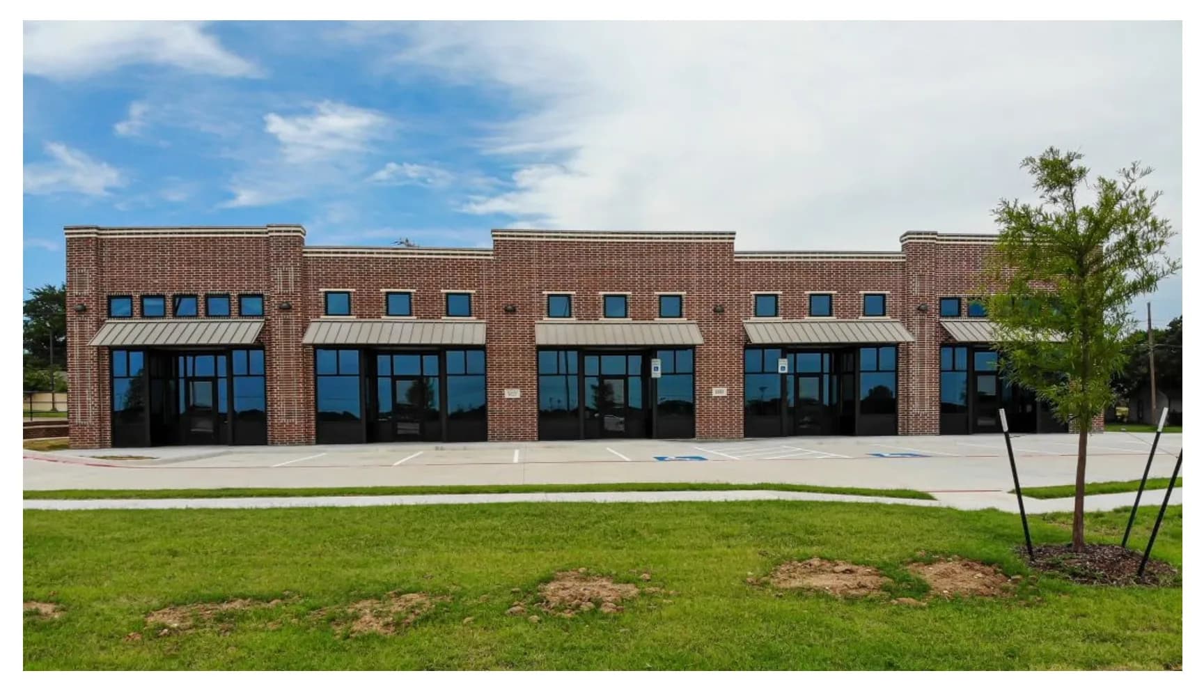 Single-story brick retail building with metal awnings, a new concrete parking lot, a young tree and fresh sod.