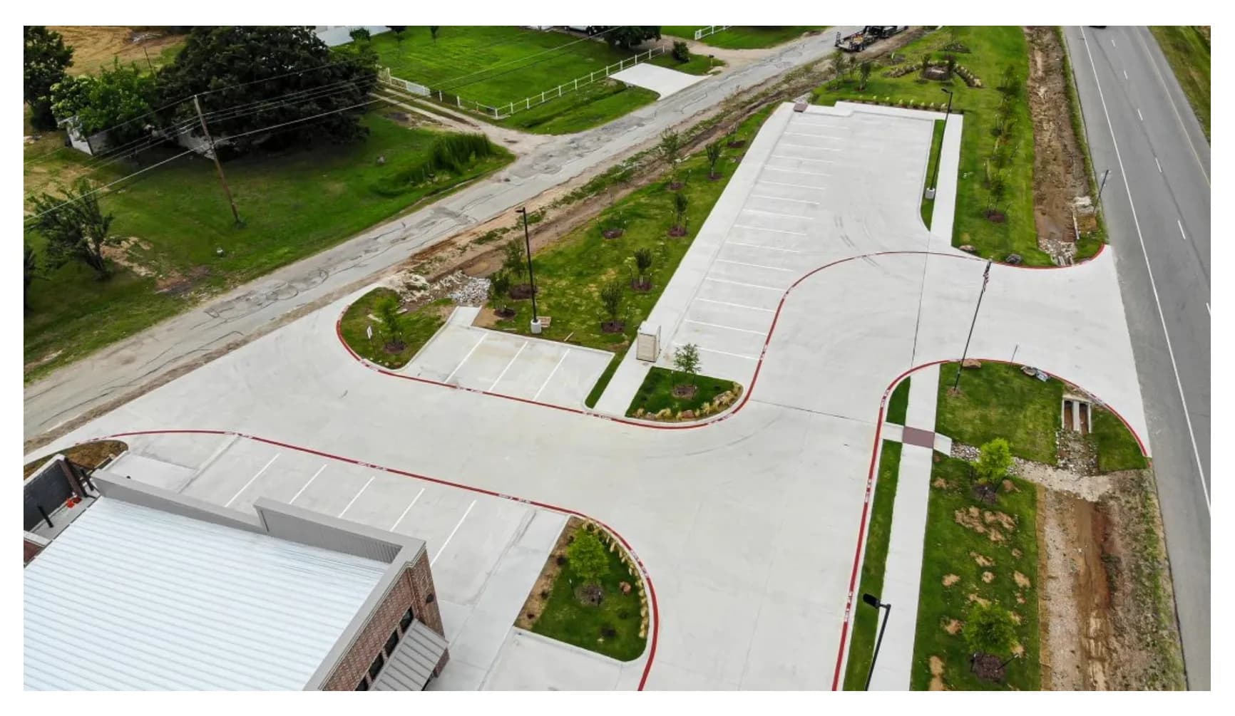 Aerial view of a finished concrete parking lot and drive with landscaped islands, light poles and a drainage swale beside a highway