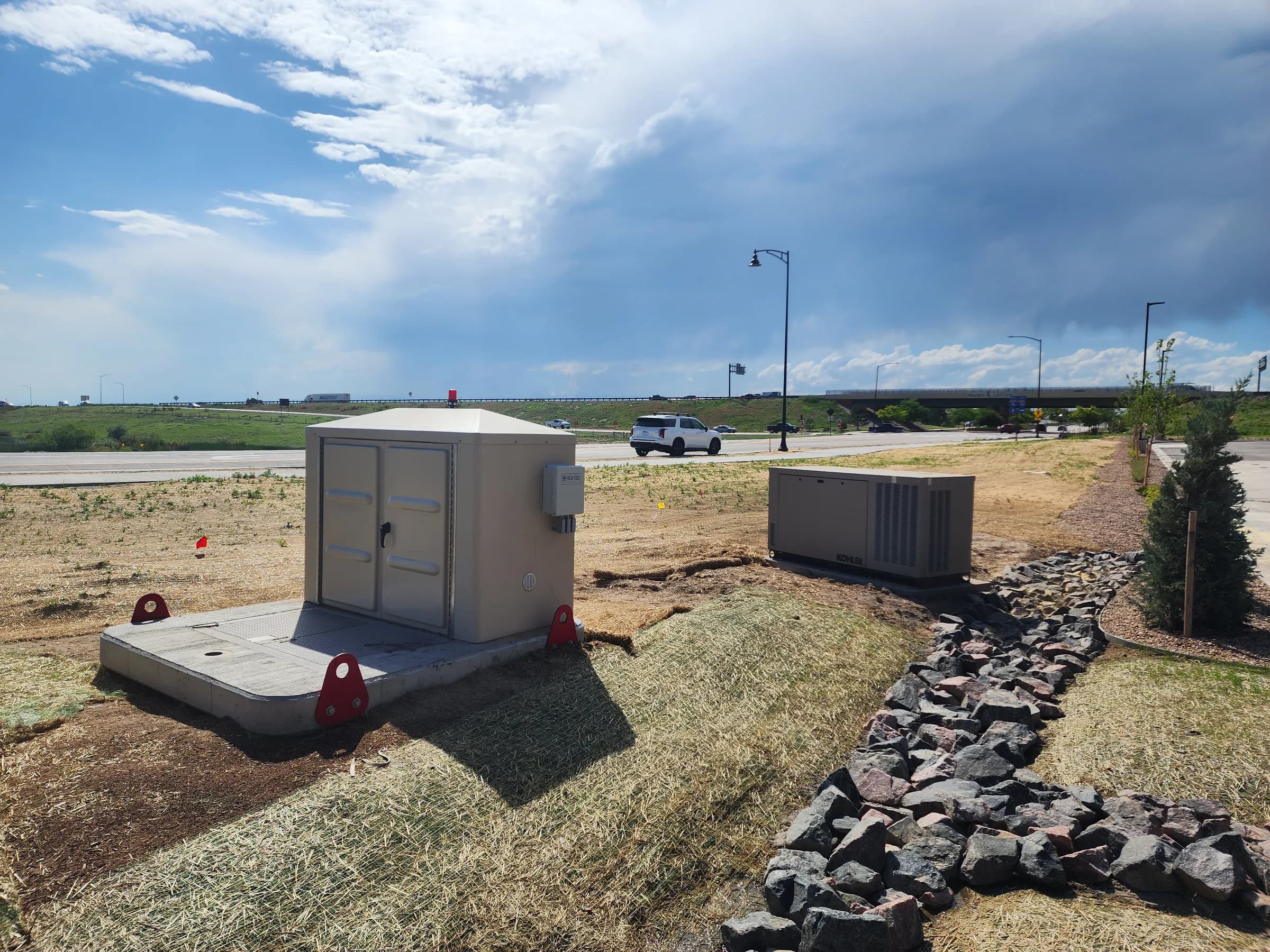A finished lift station enclosure on a concrete pad beside a standby generator and a rock-lined swale, with a highway beyond.