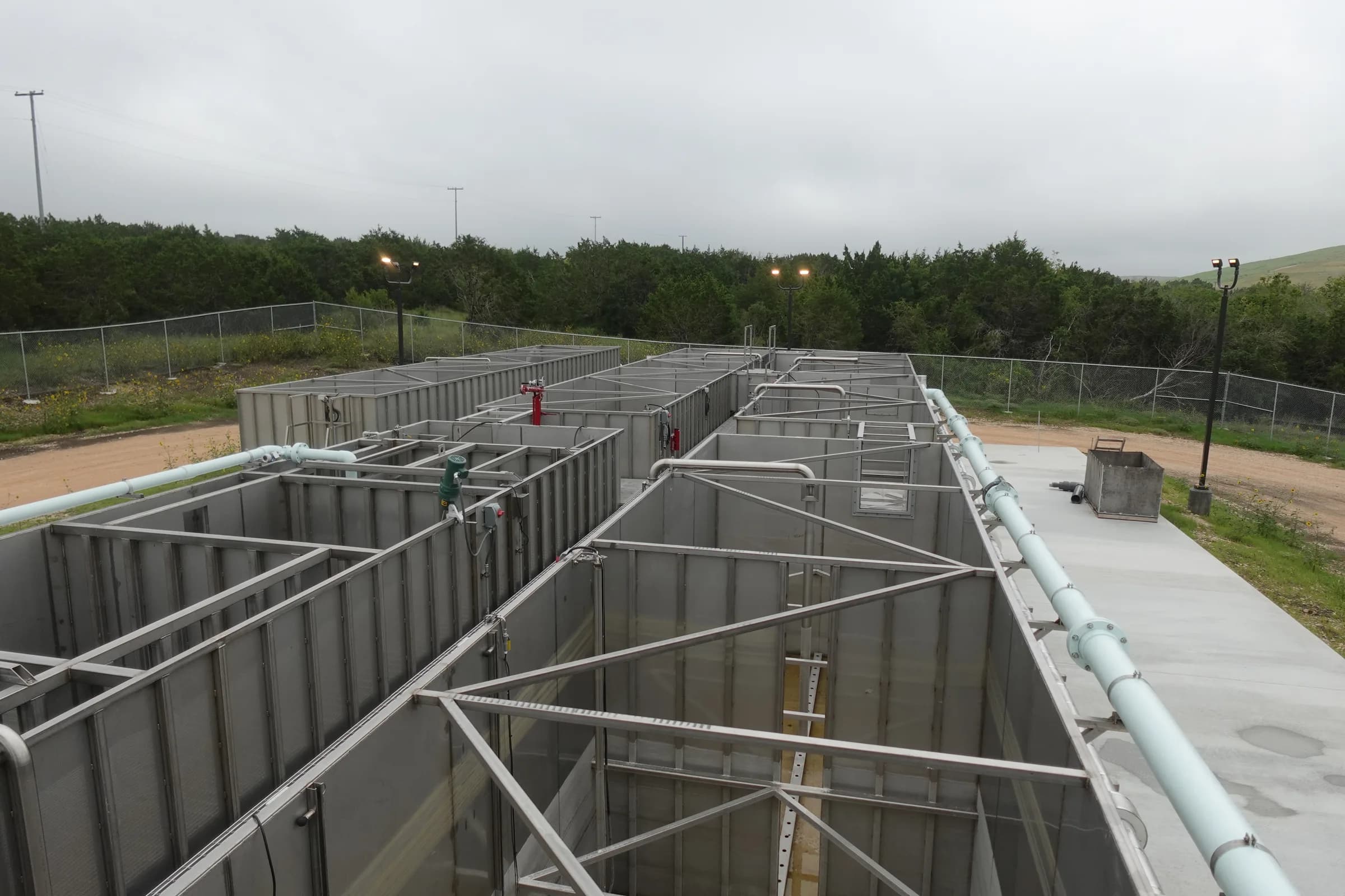 Elevated view of open steel package treatment basins with walkways and pale blue piping on a fenced concrete pad.