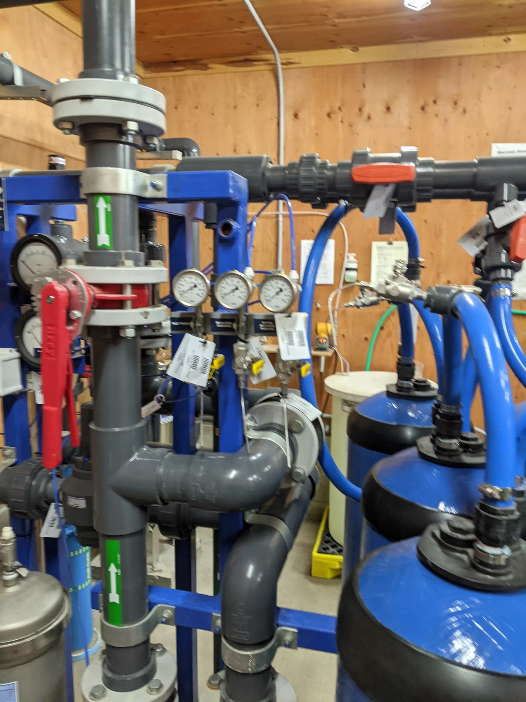 Inside a small water treatment building: gray PVC piping and pressure gauges on a blue steel frame beside blue treatment vessels
