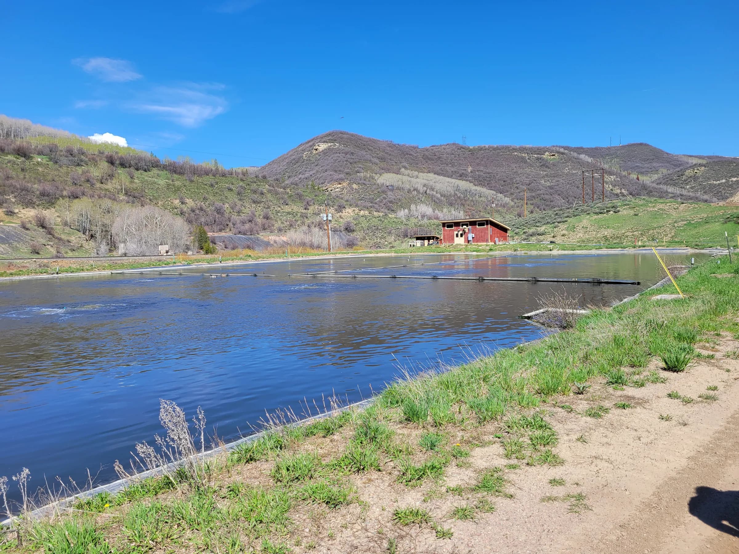 Lined wastewater lagoon beside a dirt access road, with a small red building and hills under a clear sky