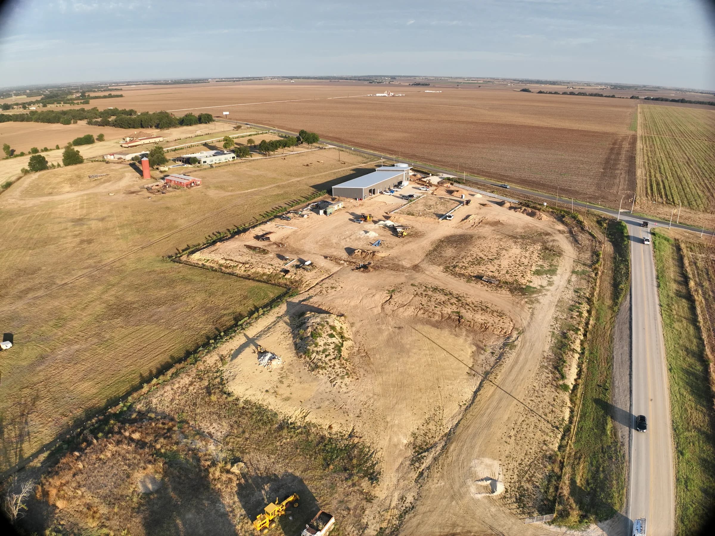 Aerial view at dusk of a graded rural site with a metal building and construction equipment beside a two-lane road and open fields.