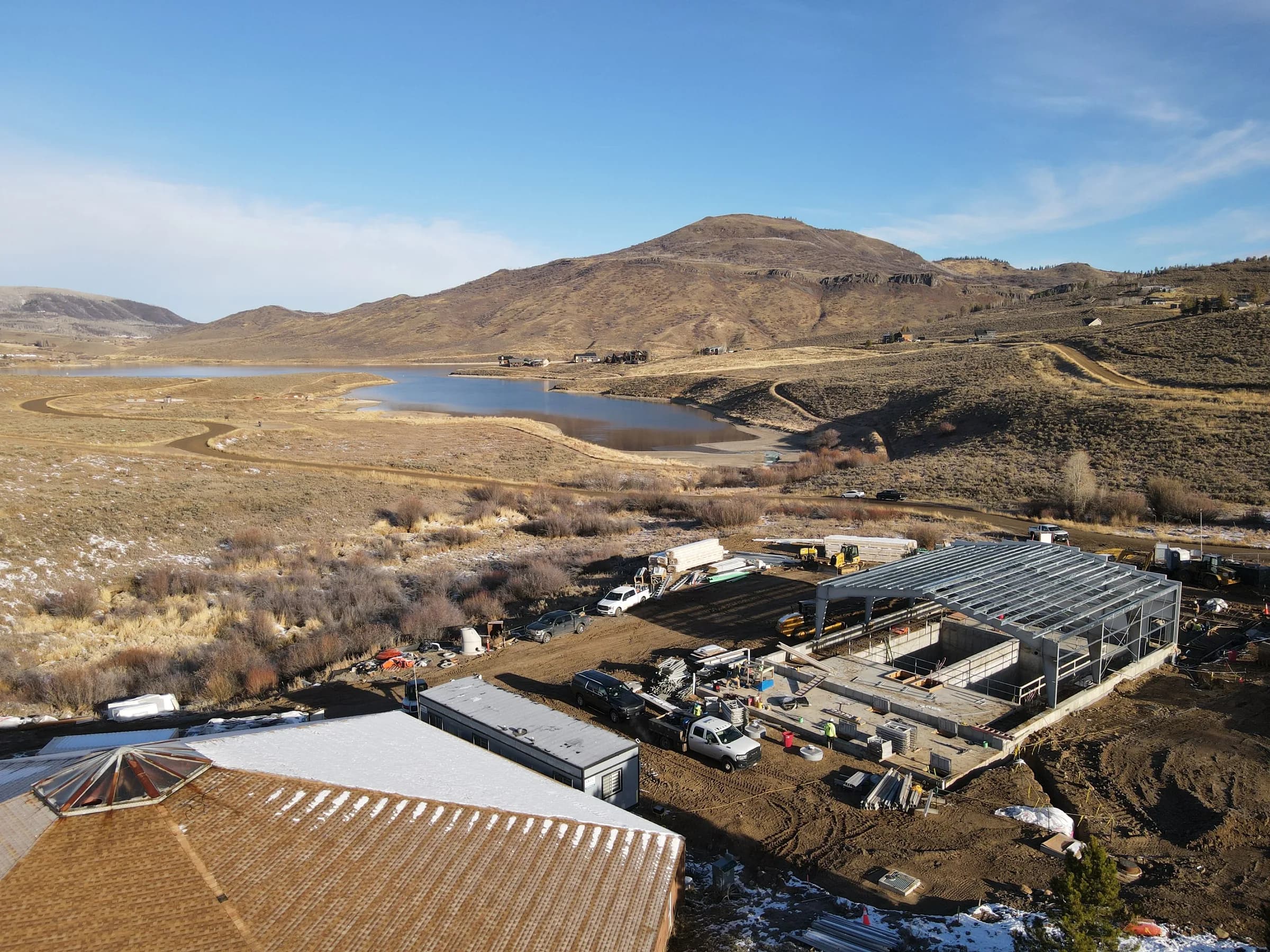 Aerial view of a wastewater treatment facility under construction beside a reservoir, with concrete basins, a steel roof frame and surrounding hills.