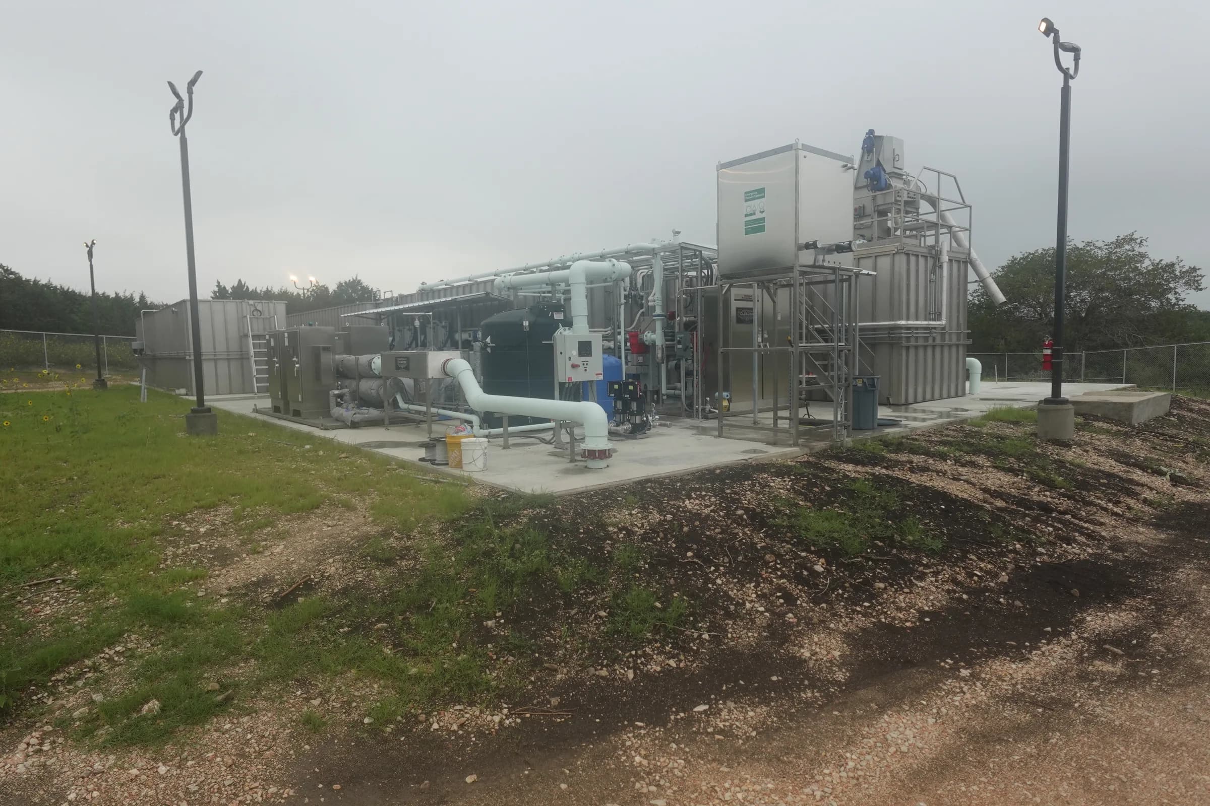 A packaged wastewater treatment plant on a concrete pad with white process piping and stainless tanks under an overcast sky.