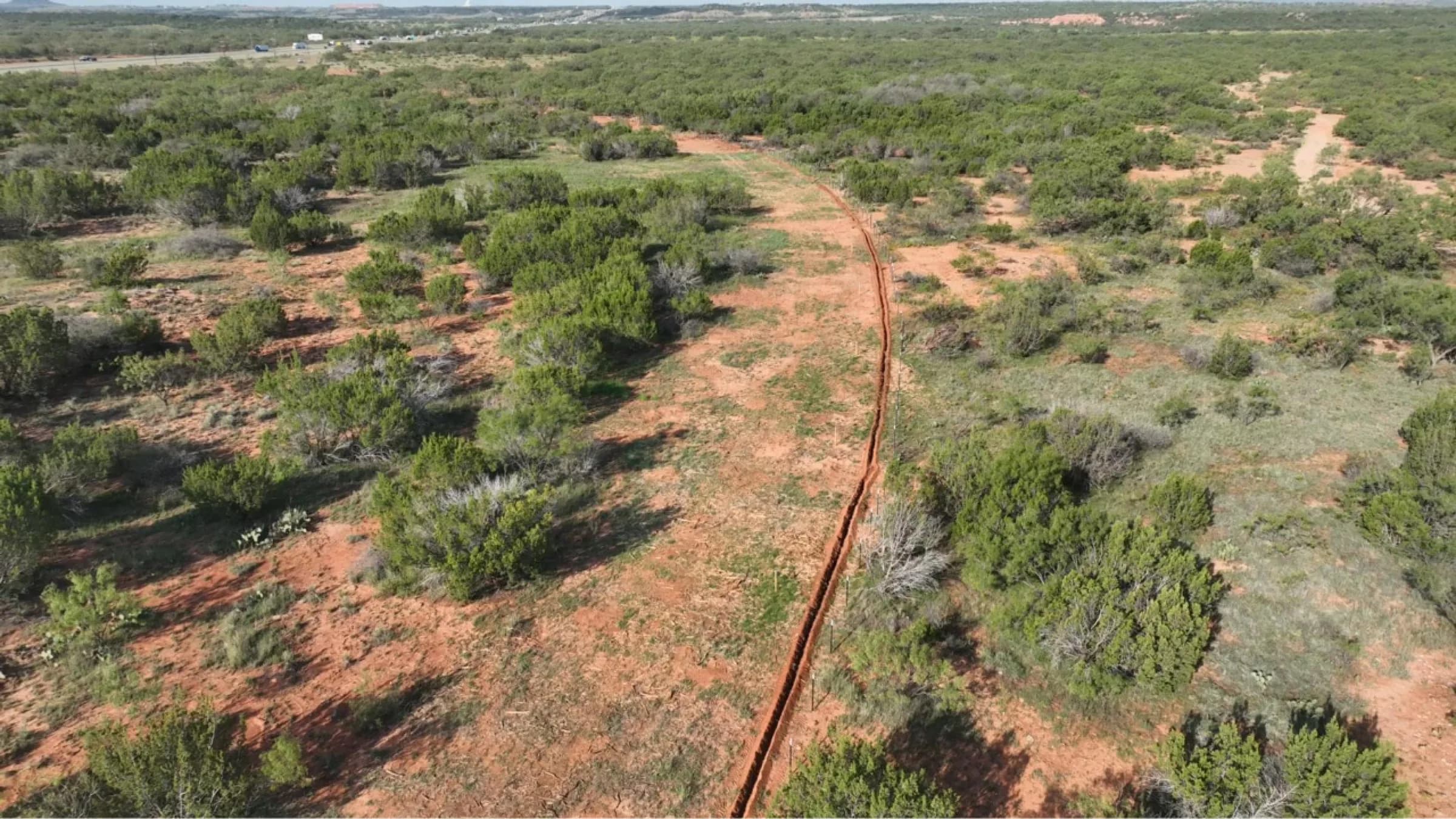 Low aerial view of a cleared trench line curving through red soil and green brush.