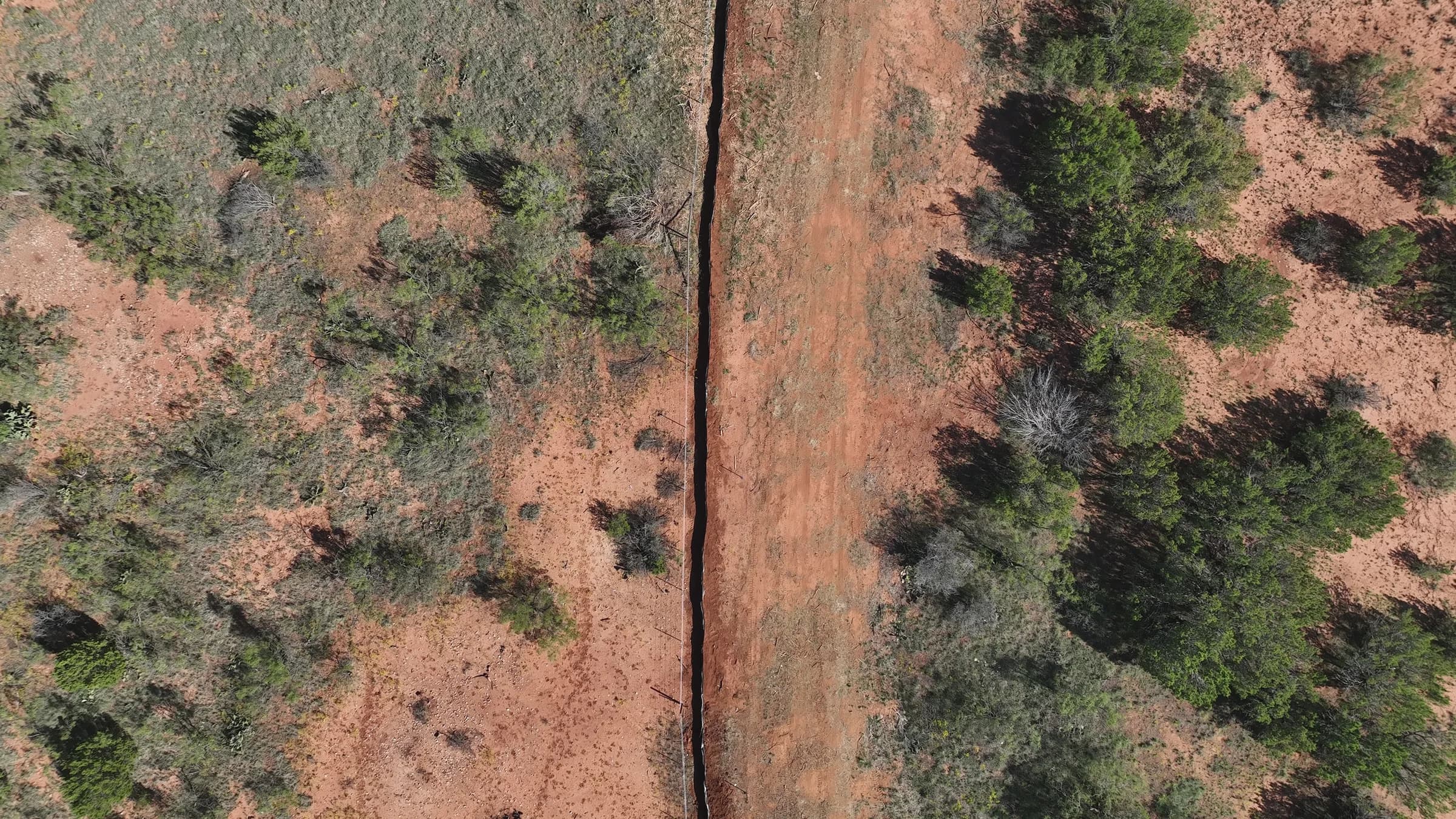 Top-down drone view of an open trench cut straight through dry brushland beside a cleared red-dirt corridor.