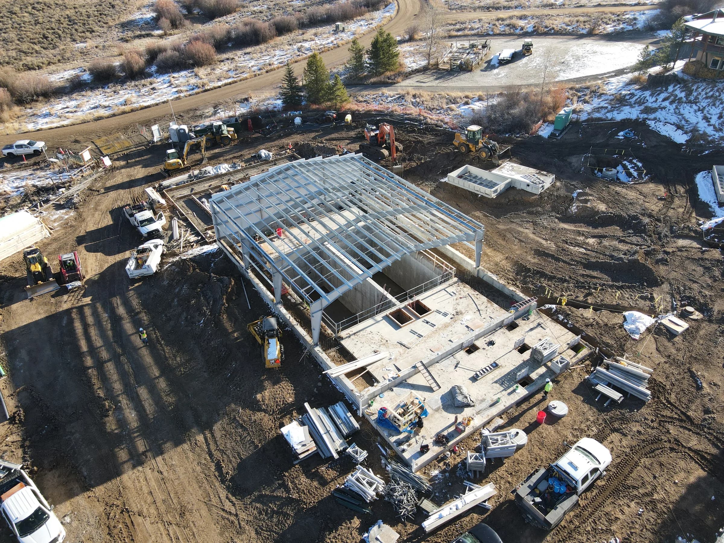 Drone view of a steel building frame rising over concrete treatment basins, with trucks and equipment around it.