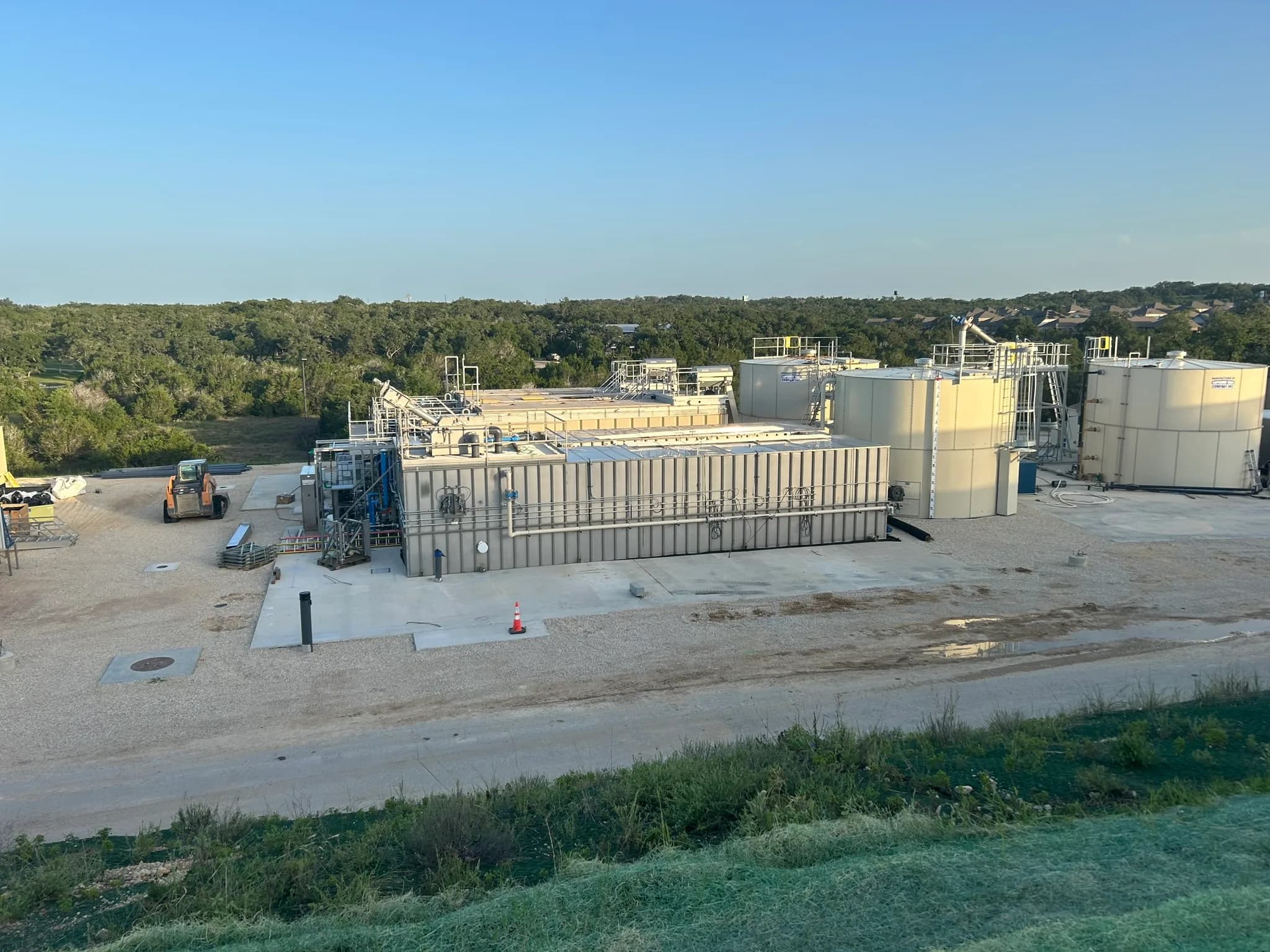 A packaged wastewater treatment plant and its tall cream tanks on a gravel pad, with trees behind.