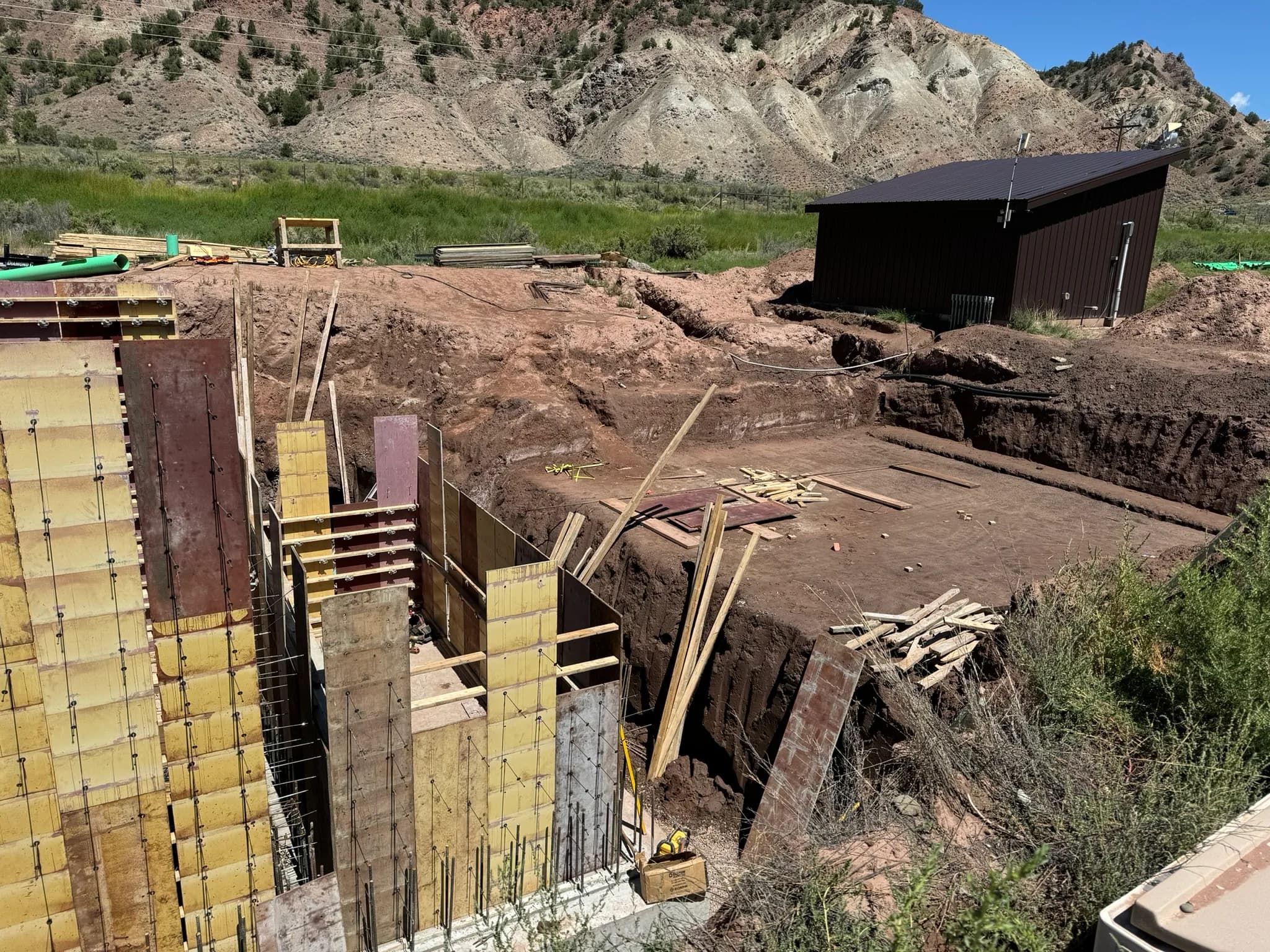 Concrete wall forms and rebar beside a deep excavation, with a small dark metal building and dry hills behind.