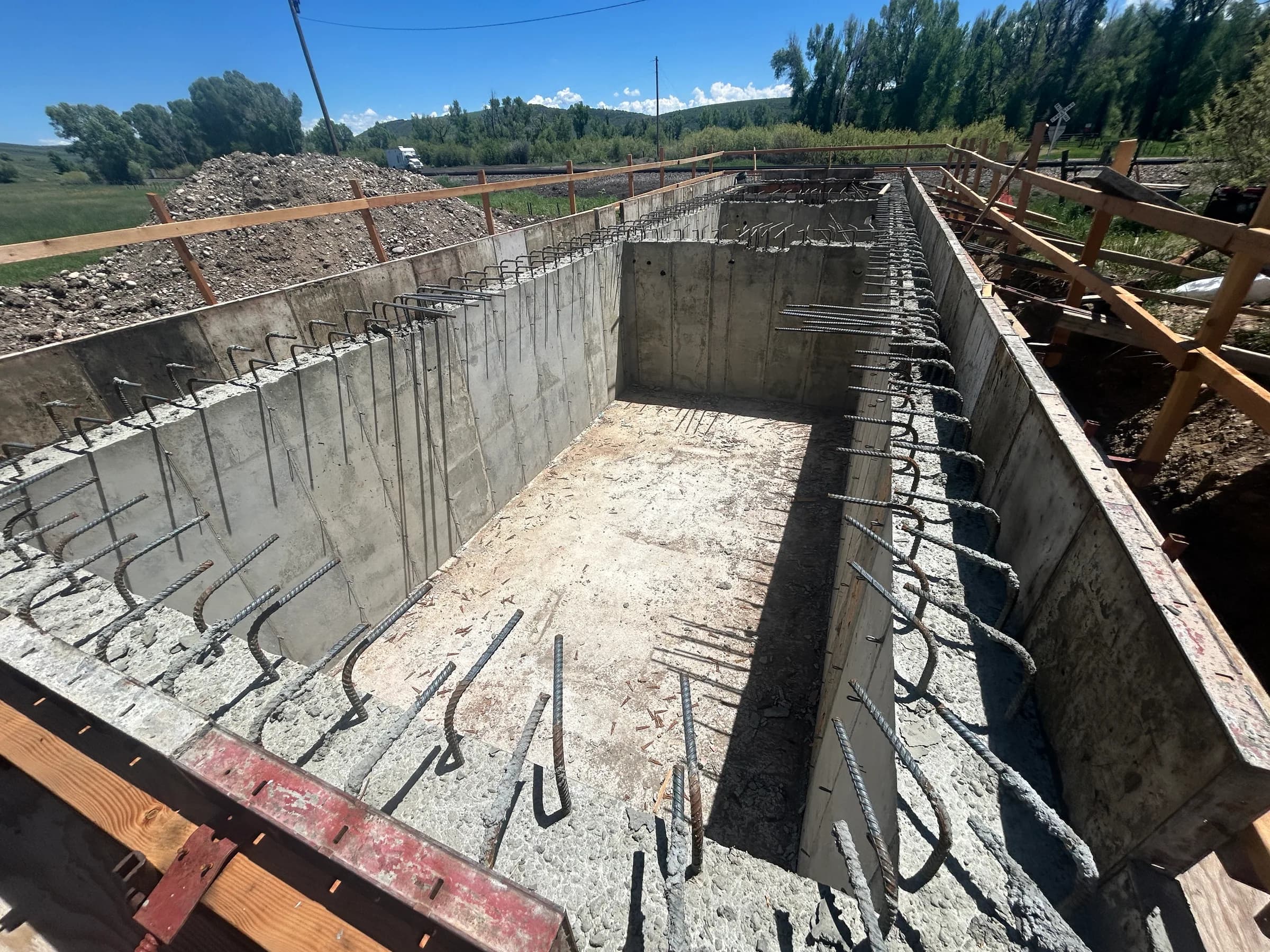 Cast-in-place concrete basin walls with rebar dowels and timber formwork on a rural construction site.