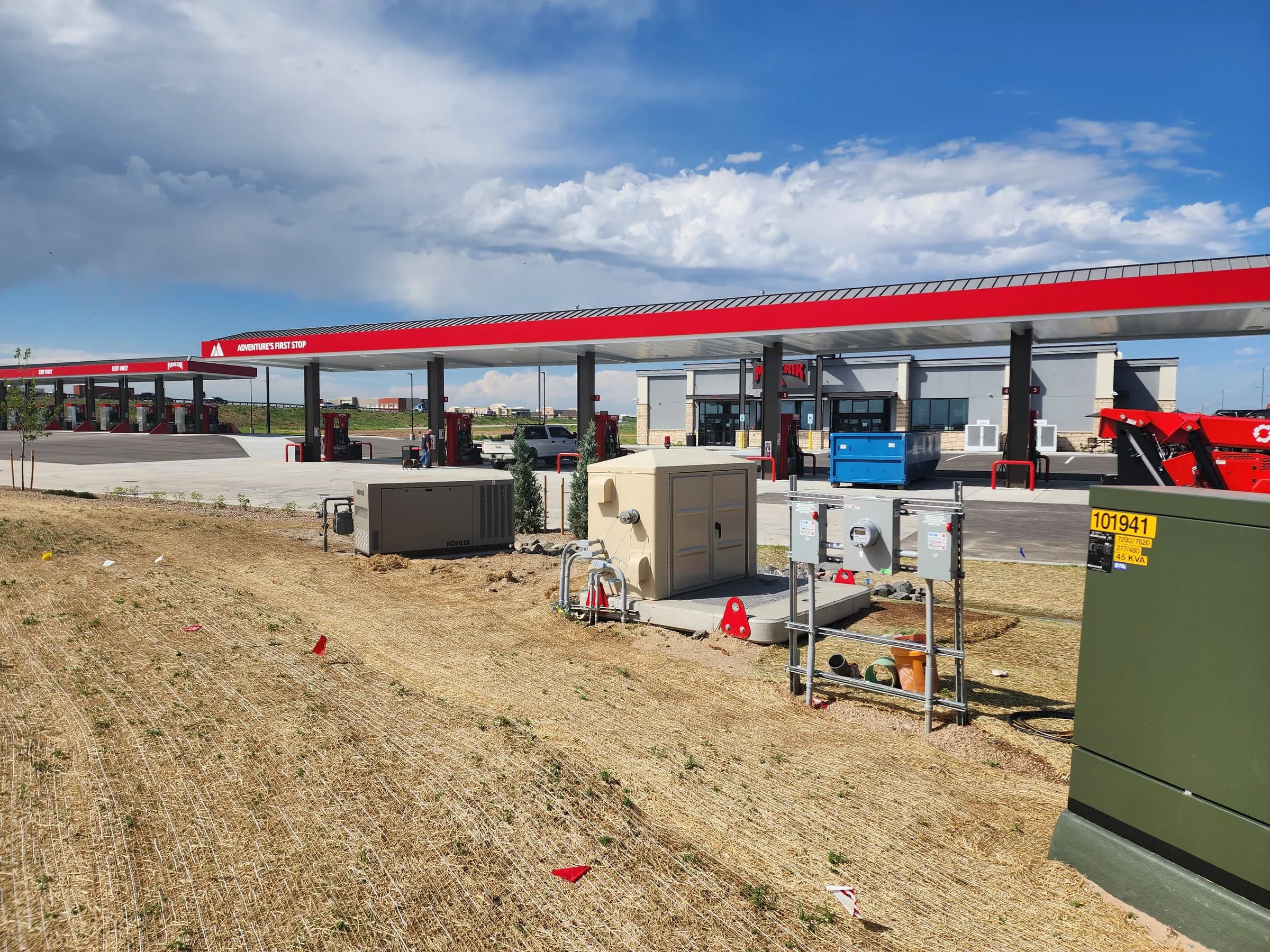 Small equipment enclosures, a generator and a utility meter rack on concrete pads in new seeded ground beside a fuel station canopy.
