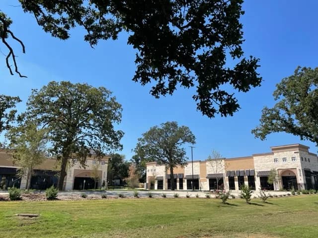 Finished stone and brick commercial buildings with landscaped grounds and mature oak trees under a clear blue sky.