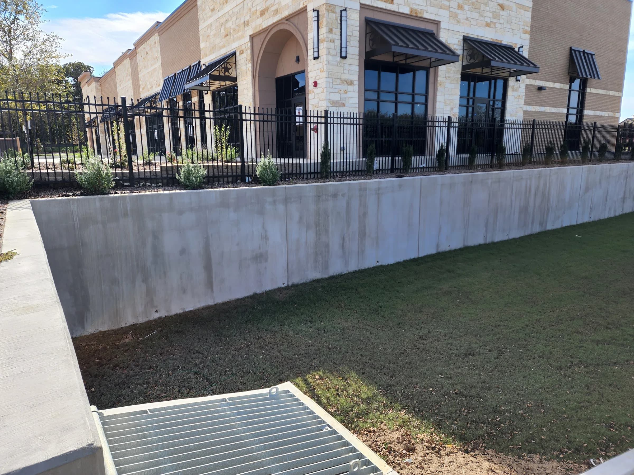 Grass-lined drainage channel with a concrete retaining wall and galvanized grate inlet beside a new stone and brick commercial building.