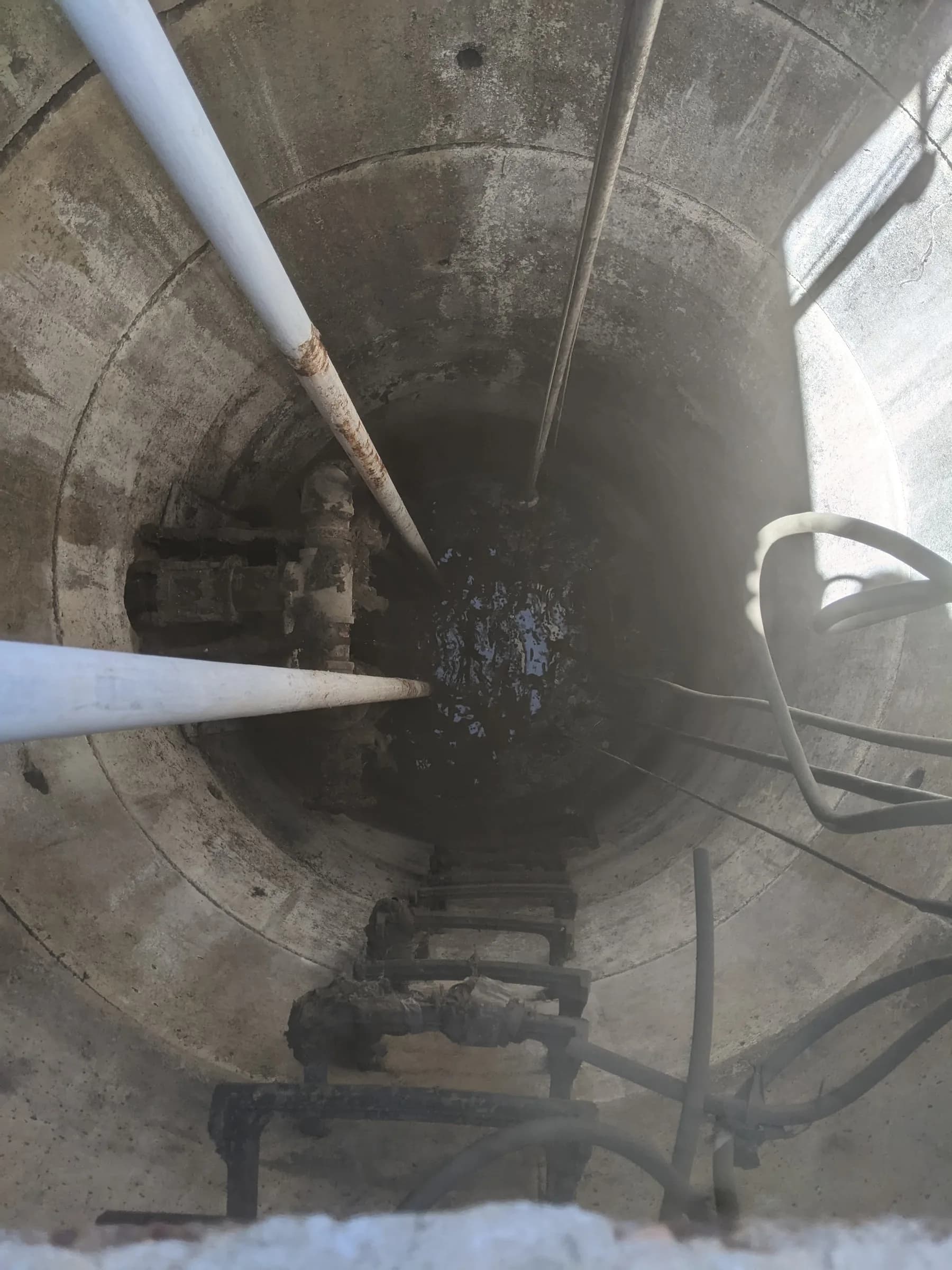 Looking down into a concrete lift station wet well with guide rails, discharge piping, a ladder and hoses above standing water