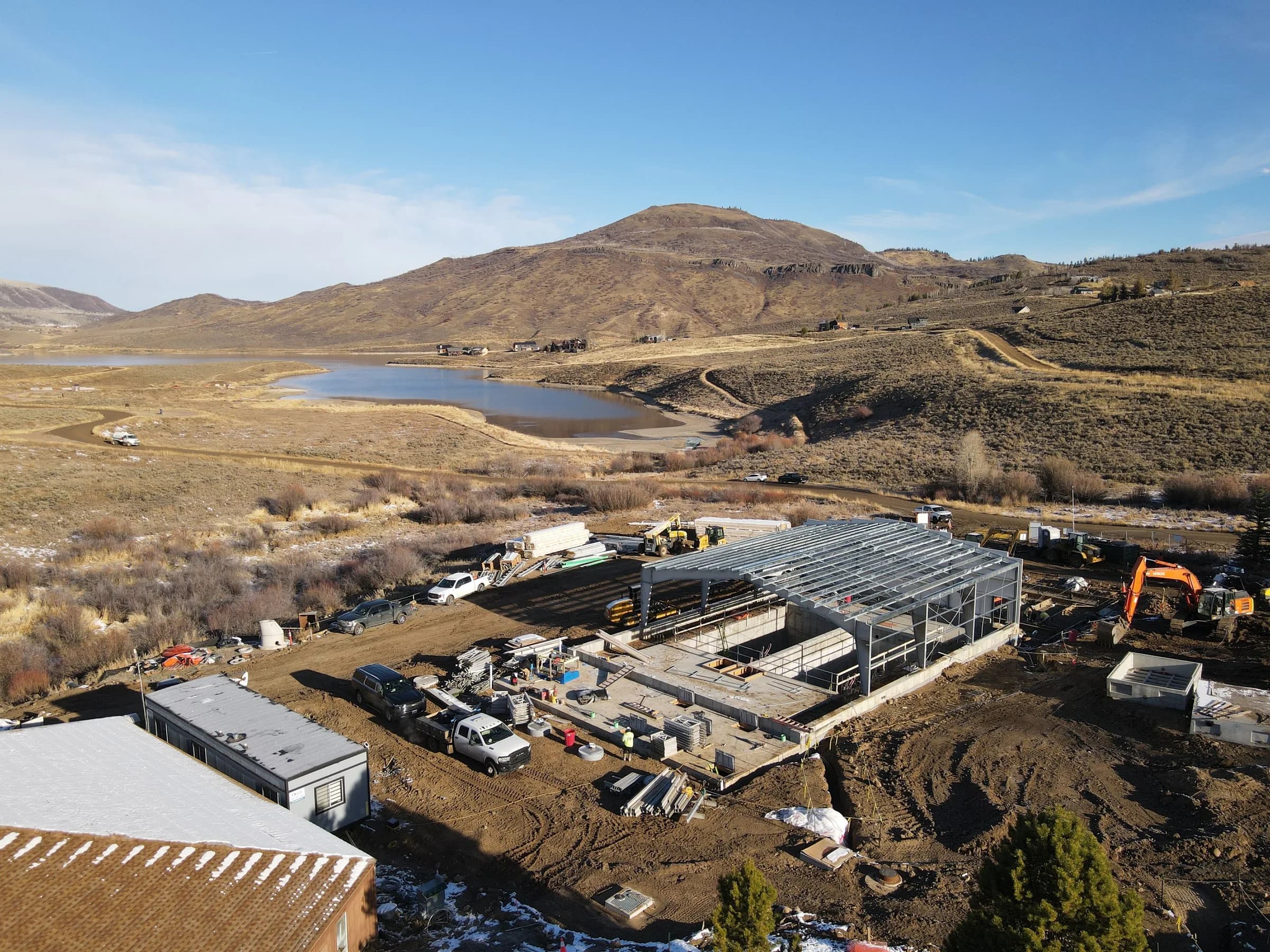 Aerial view of a steel-framed treatment building going up on a muddy site with an excavator and trucks.