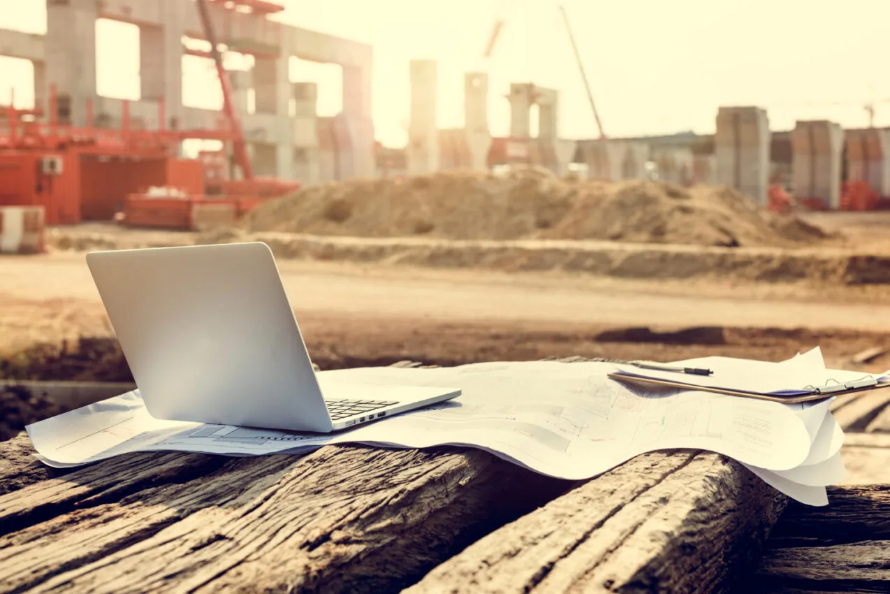 Laptop and construction drawings on a weathered timber beam at a construction site, with concrete columns, a crane and red equipment in the background.