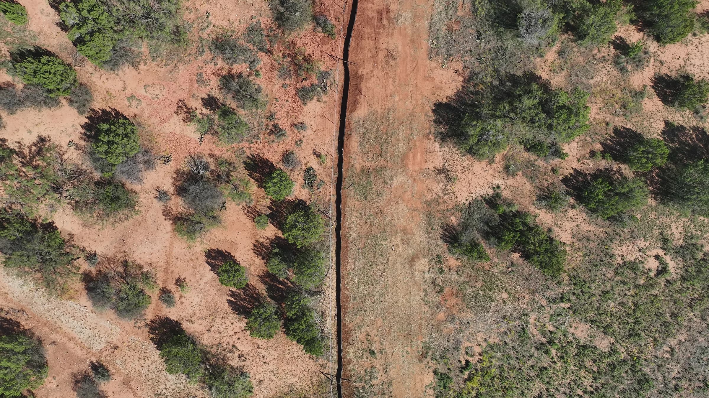Straight-down drone view of a narrow open trench cutting through red soil and green scrub.