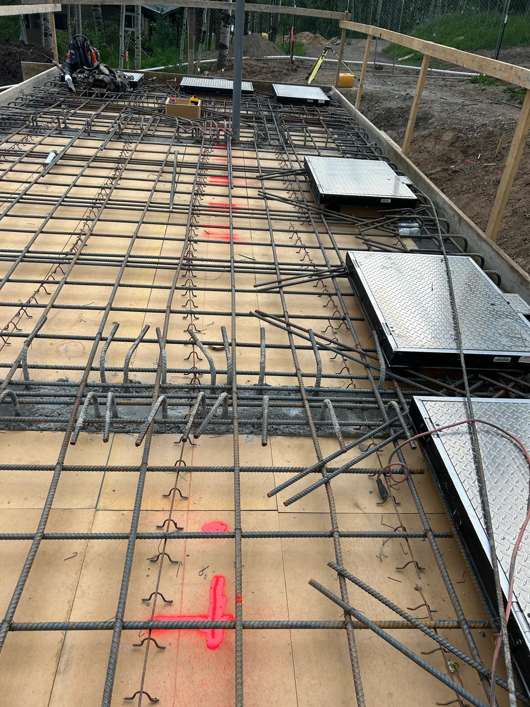 Rebar mat on a formed deck with aluminum diamond-plate access hatches set in place, with excavated ground beyond.