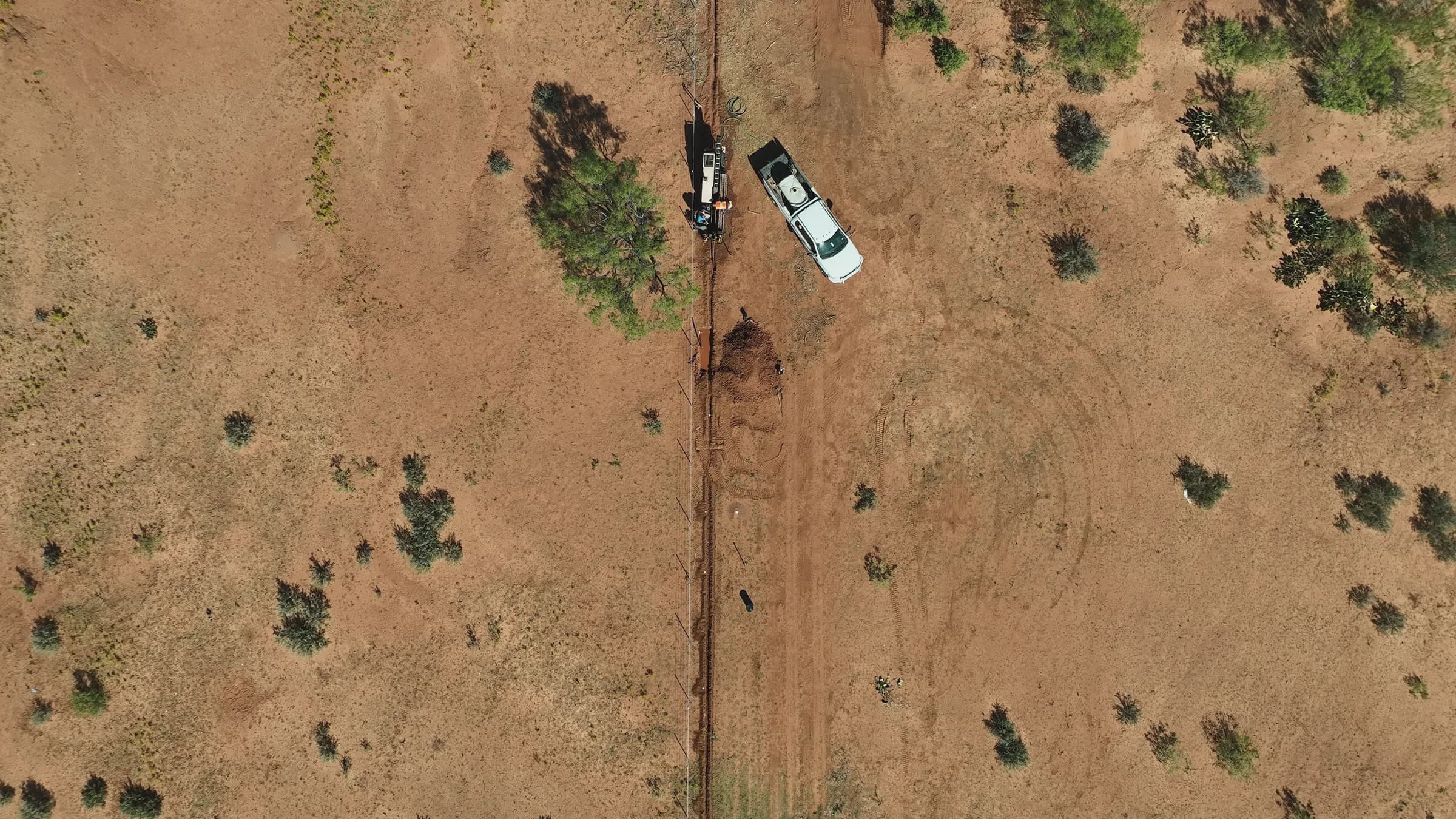 Overhead drone view of a drill rig truck and a pickup beside an open trench in dry scrubland.