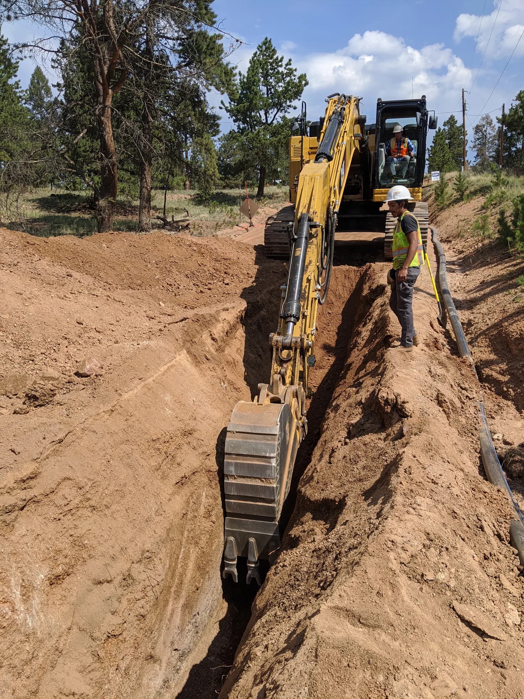 Project photo: an excavator digs a utility pipeline trench while a crew member watches.