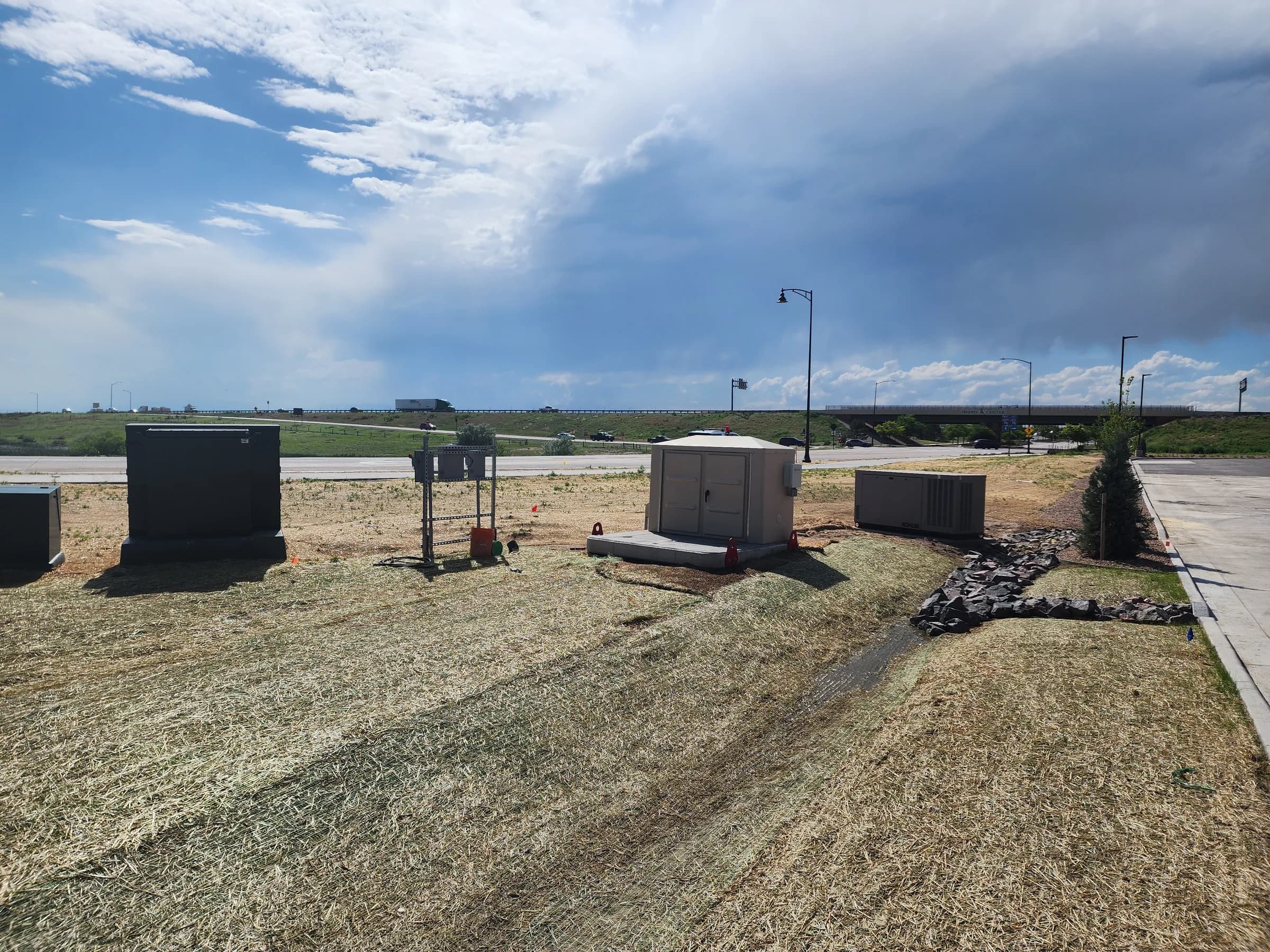 Lift station control cabinets and a backup generator beside a rock-lined drainage swale on newly seeded ground near a highway interchange