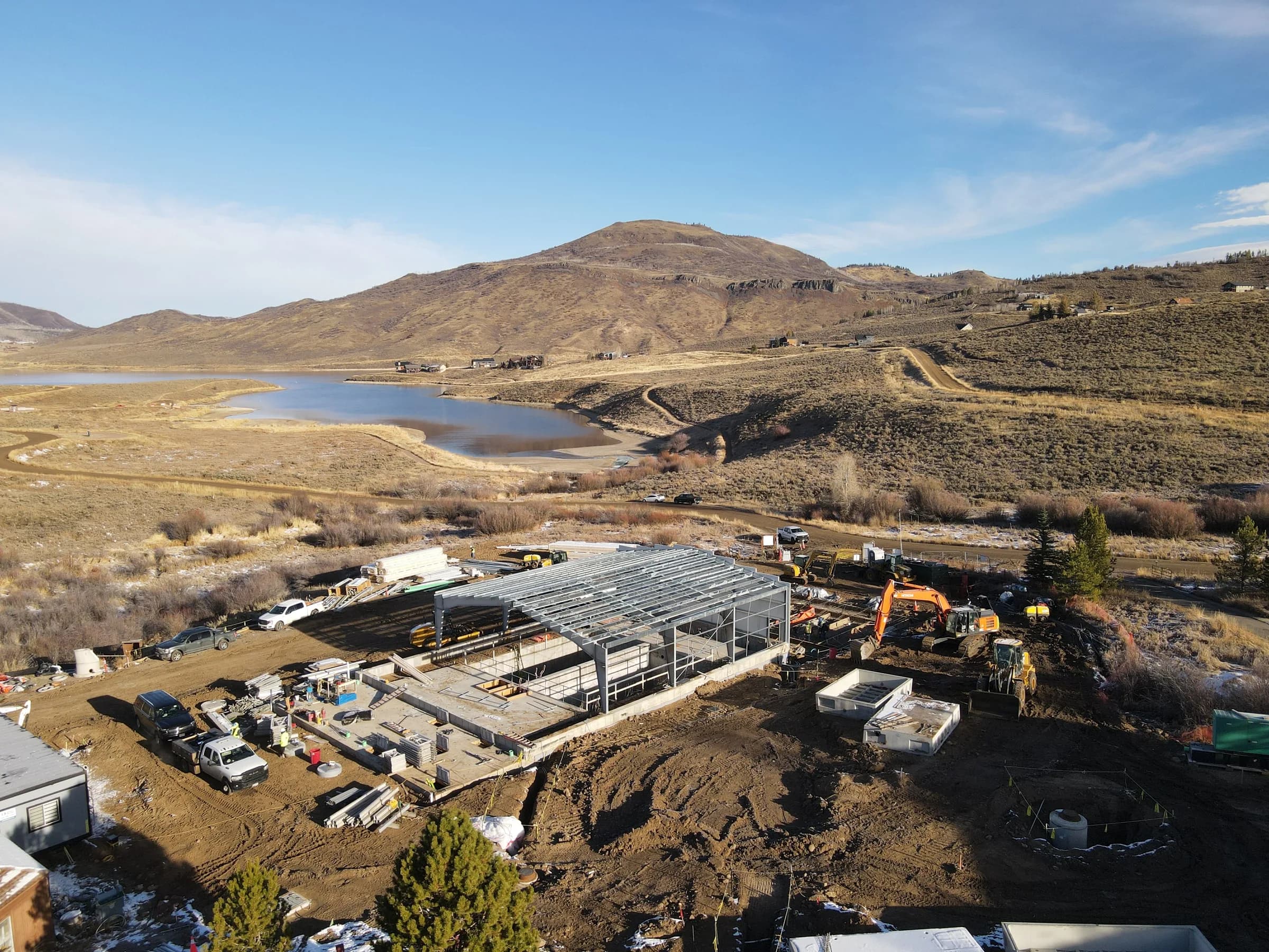 Drone view of a new treatment building’s steel frame on its slab, with excavators and site work around it and a reservoir and a mountain behind.