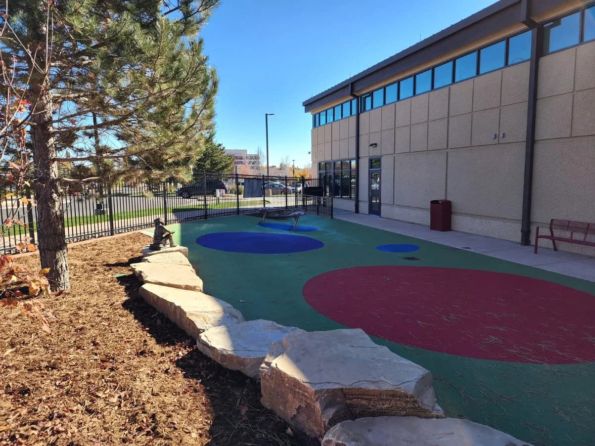 Finished outdoor play area with colorful rubber surfacing, boulder seating and mulched landscaping beside a building