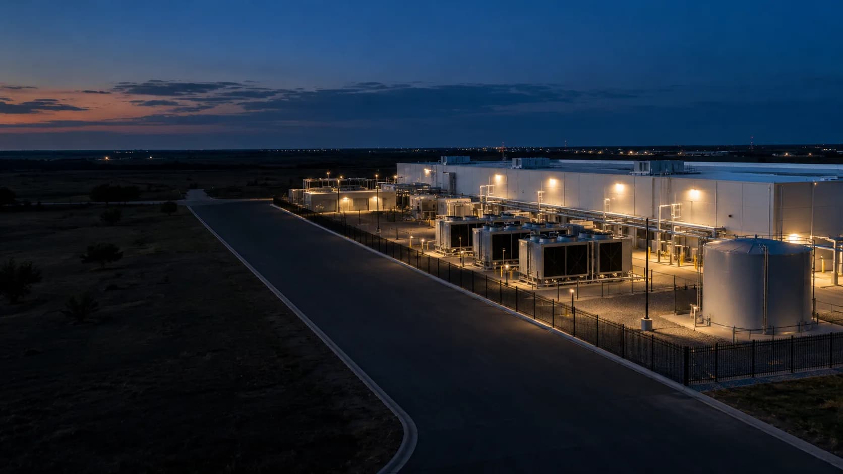 Aerial dusk view of a large industrial facility with rooftop units, cooling equipment, a water storage tank, fenced yard and paved access road.