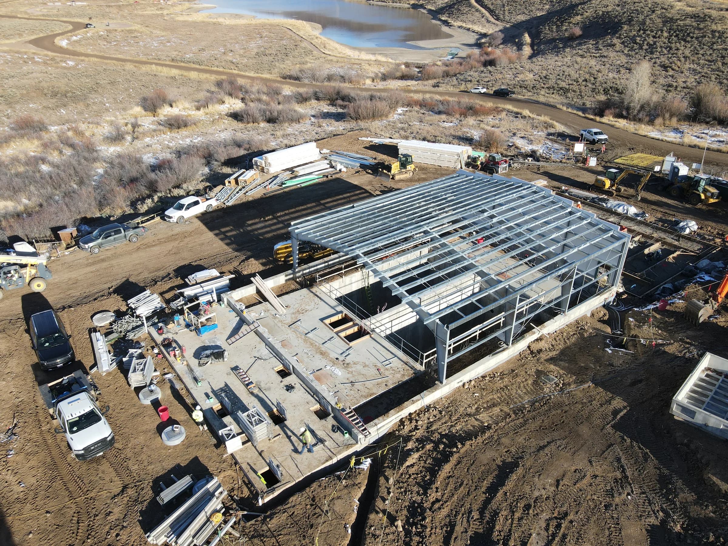 Drone view of a treatment plant under construction, with a steel roof frame over concrete basins and a reservoir beyond