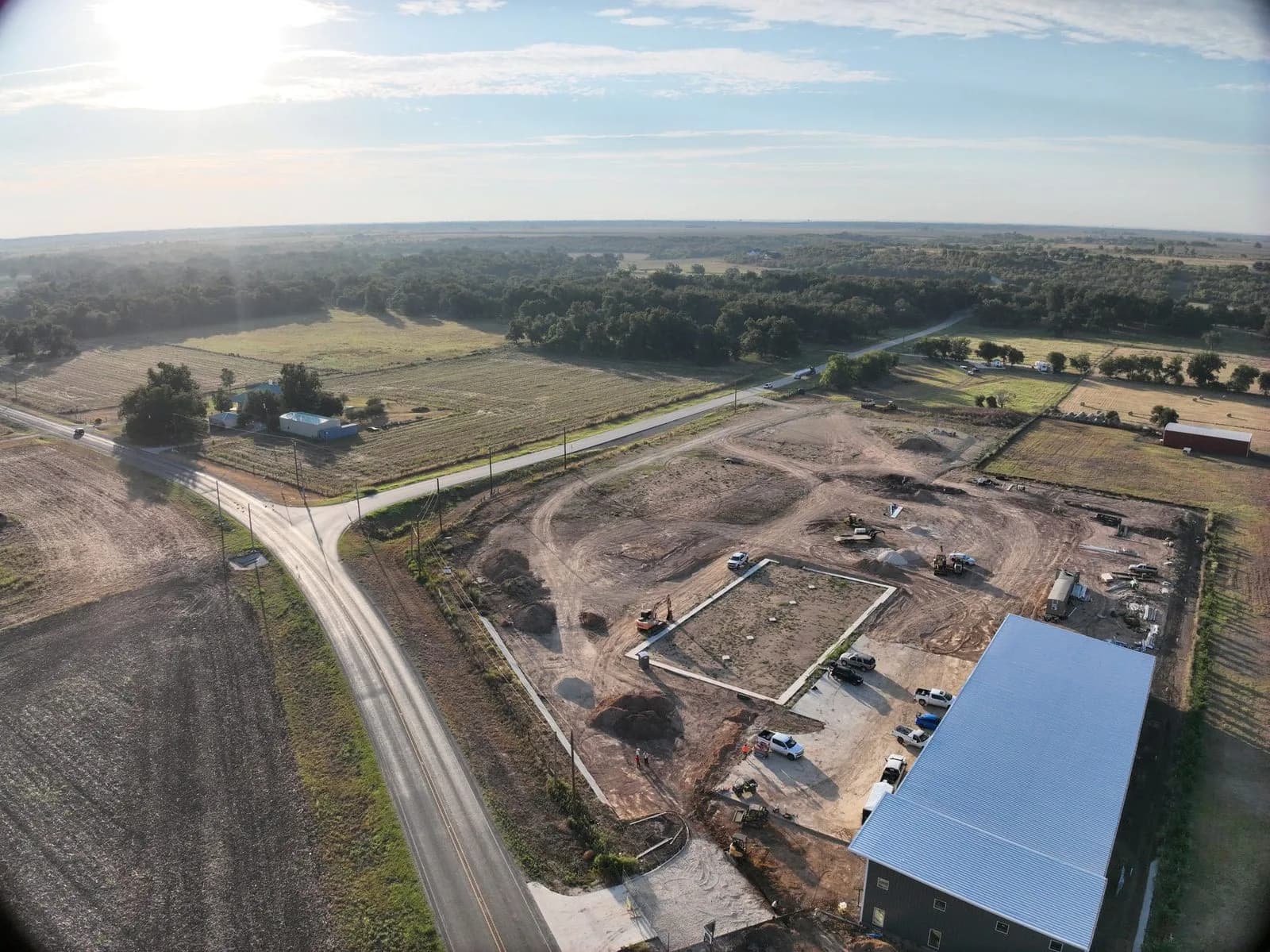 Wide aerial view of a rural commercial site under construction, with a new metal-roofed building and a graded pad.