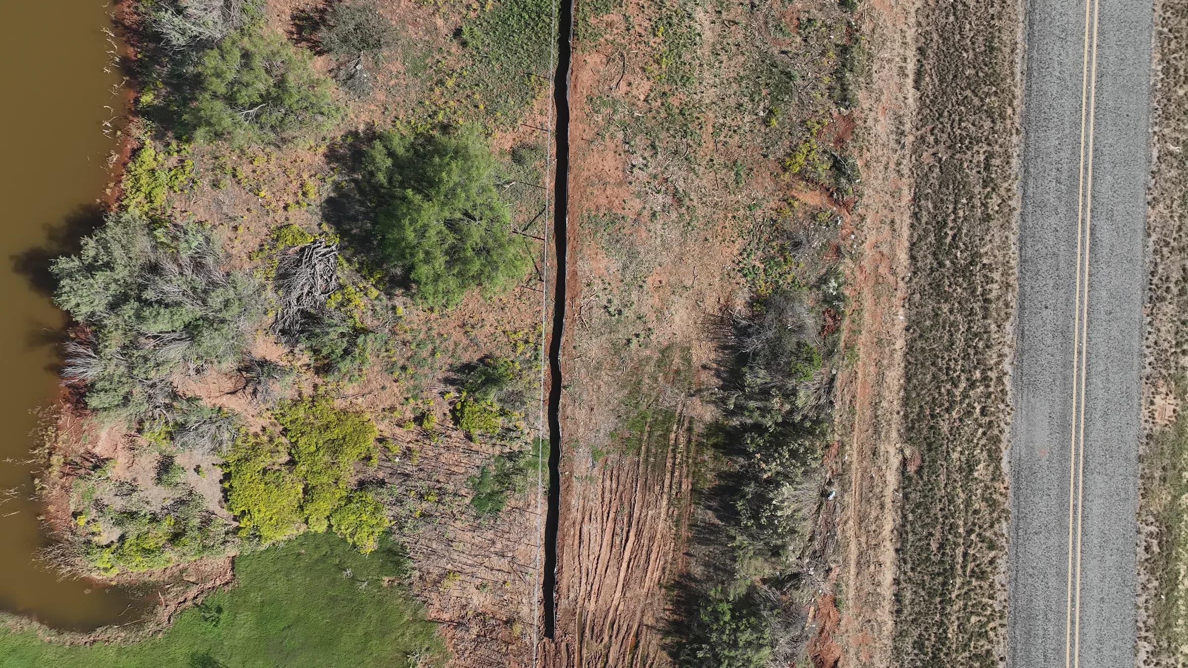 Overhead drone view of a long open trench through brush between a pond and a paved two-lane road.