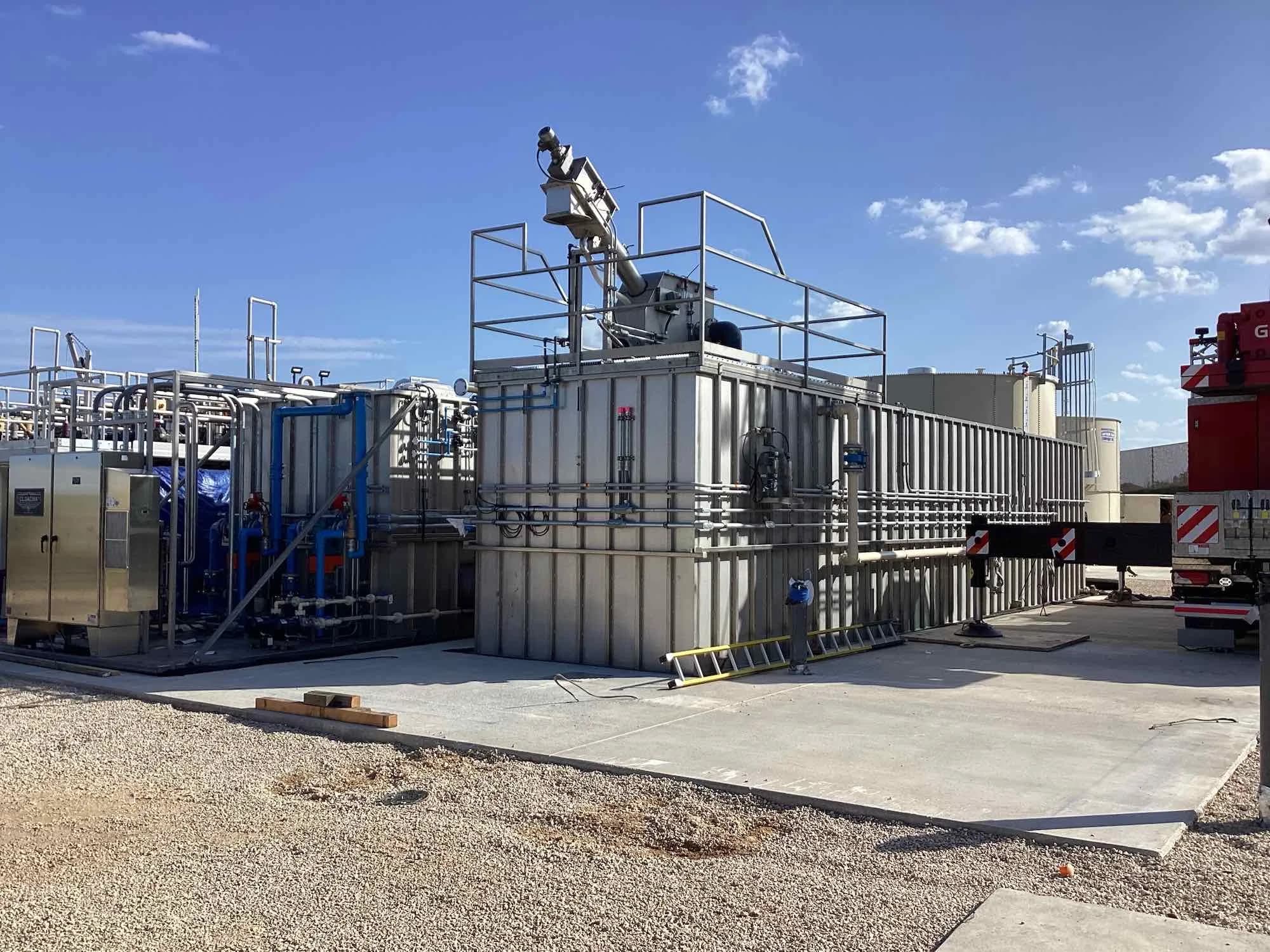 Packaged wastewater treatment unit with a screen and railed platform on a concrete pad, with piping and tanks nearby.