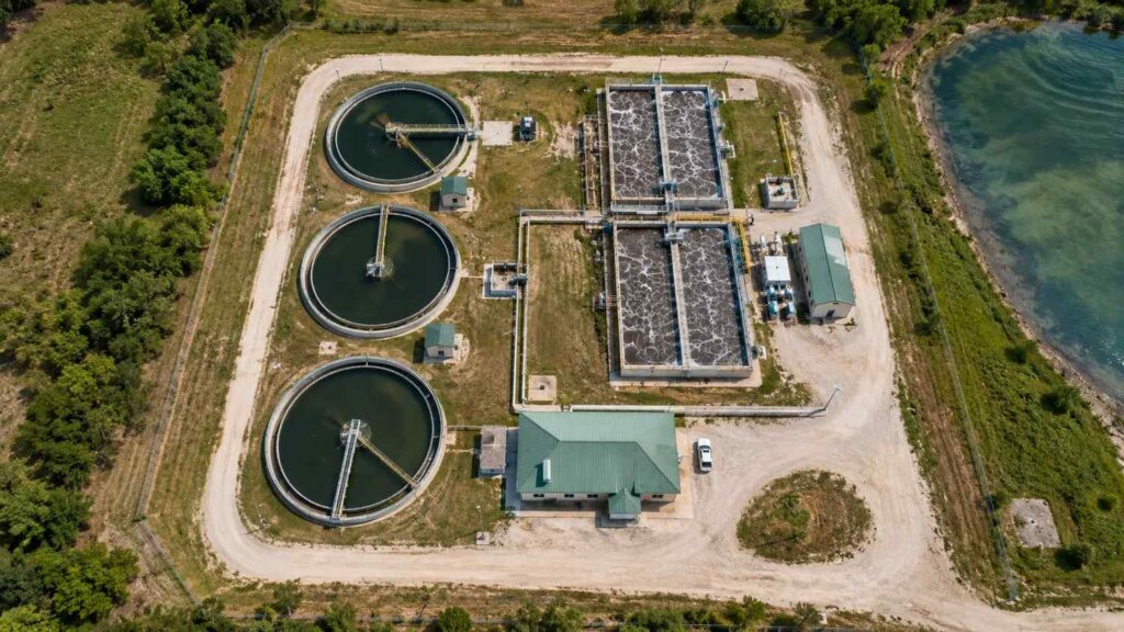 Top-down aerial view of a wastewater treatment plant showing multiple circular clarifier tanks, aeration basins, and treatment process infrastructure from above with engineering modeling equipment visible at an operations building