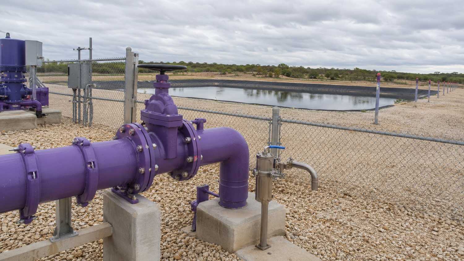 Texas reclaimed water distribution system showing purple painted exposed piping at a pump station connection point with a lined reclaimed water storage pond visible in the background and sampling port equipment at the distribution system outlet