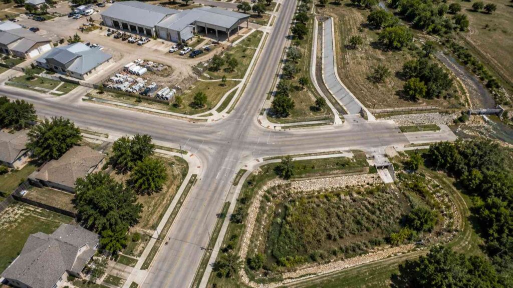 Top-down aerial view of a small Texas city showing storm sewer infrastructure including street drainage inlets, outfall structures discharging to a creek, and municipal facilities representing the regulated MS4 system requiring Phase II permit compliance