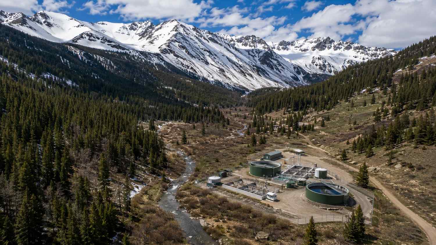 Aerial view of a Colorado mountain watershed showing spring snowpack on high ridgelines above a valley with a small stream and wastewater treatment facility representing the seasonal hydrologic cycle that drives CDPHE discharge permit conditions evaluated by Modern Engineering Solutions.