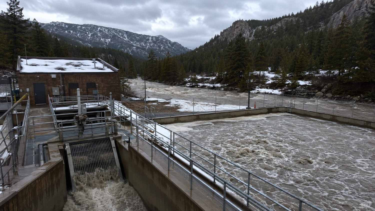 Ground-level shot of a Colorado mountain wastewater treatment plant during spring snowmelt showing elevated influent flows entering the headworks with visible high water levels in aeration basins representing the infiltration and inflow hydraulic overloading challenge that Modern Engineering Solutions addresses through collection system evaluation and flow equalization design.