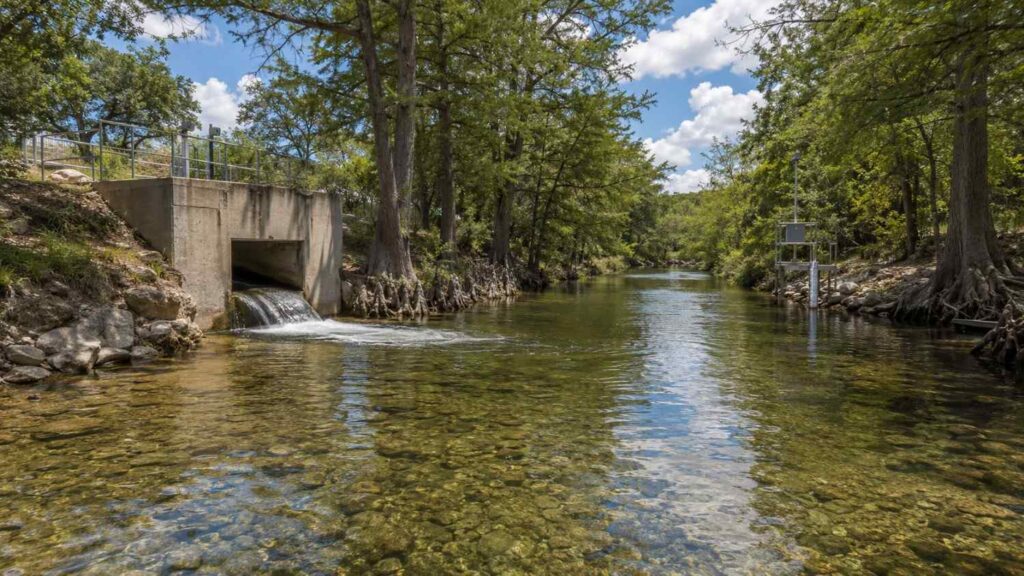 Ground-level wide shot of a treated effluent outfall discharging to a clear Central Texas Hill Country creek surrounded by cypress trees and limestone banks representing the receiving water quality standards and designated use classifications that drive TCEQ effluent limit calculations in TPDES permits that Modern Engineering Solutions evaluates for Texas discharge projects