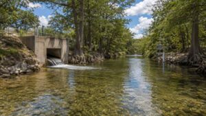 Ground-level wide shot of a treated effluent outfall discharging to a clear Central Texas Hill Country creek surrounded by cypress trees and limestone banks representing the receiving water quality standards and designated use classifications that drive TCEQ effluent limit calculations in TPDES permits that Modern Engineering Solutions evaluates for Texas discharge projects