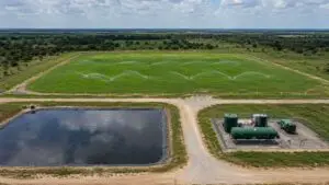 Top-down aerial view of a Texas land application site showing active spray irrigation system covering a large green pasture field with a wastewater treatment facility visible at the site edge and a lined evaporation pond in the background representing the three primary TLAP disposal methods